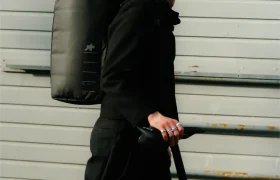 Una persona vestida con ropa negra, gorra y gafas de sol se sitúa junto a una bicicleta con una gran mochila negra ASSOS x Db, encarnando el espíritu del sistema de viaje para ciclistas contra una pared metálica de color claro.