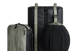 Un juego de bolsas de viaje negras y grises de la colección ASSOS x Db, diseñado para ciclistas, con un cubo de embalaje rectangular y una bolsa de viaje cilíndrica, con asas resistentes y un inteligente sistema de viaje.