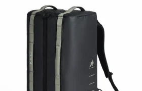 Una mochila rectangular negra con correas grises, un asa superior y una única correa para el hombro, diseñada para ciclistas como parte del sistema de viaje ASSOS x Db, en posición vertical sobre fondo blanco.