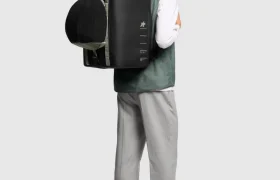 Persona con gorra verde, chaleco verde, camisa blanca, pantalón gris y zapatos beige, que lleva una mochila negra rectangular grande ASSOS x Db, parte de un innovador sistema de viaje ideal para ciclistas.