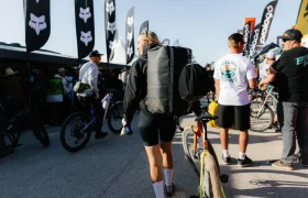 Una persona con una gran mochila ASSOS x Db y una bicicleta camina a través de un concurrido evento al aire libre lleno de ciclistas, bicicletas, banderas y gente con atuendo informal.