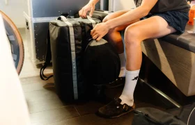 Una persona en calcetines y sandalias está sentada en el interior, cerrando la cremallera de una gran bolsa negra ASSOS x Db en el suelo: un sistema de viaje ideal para los ciclistas que se preparan para su próxima aventura.