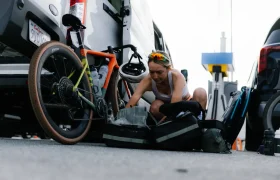 Una mujer, arrodillada junto a una furgoneta aparcada, organiza las bolsas de equipo junto a una bicicleta con neumáticos de tierra en un aparcamiento: su eficaz sistema de viaje es perfecto para ciclistas en movimiento.