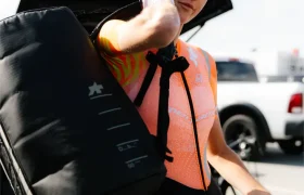 Un ciclista con casco y gafas de sol, luciendo el sistema de viaje ASSOS x Db, lleva una gran bolsa negra sobre un hombro en un aparcamiento con el maletero de un coche abierto detrás.
