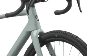 Primer plano de una bicicleta SCOTT Addict Gravel 2027 de color gris claro en el que se aprecian la rueda delantera, la cubierta con banda de rodadura de tacos, ideal para recorridos por grava, la horquilla, el manillar y las manetas de freno. Esta bicicleta polivalente está diseñada para rendir al máximo en una gran variedad de terrenos.