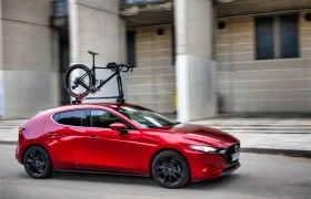 Un utilitario rojo con un portabicicletas Allen Smart Suction montado en el techo transporta una bicicleta mientras pasa por delante de un moderno edificio de hormigón.