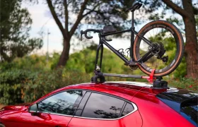 Una bicicleta está montada en un portabicicletas Allen Smart Suction Bike Rack sobre un coche rojo, aparcado al aire libre con árboles y vegetación de fondo.