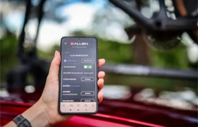 Una mano sostiene un smartphone que muestra la interfaz de la aplicación de Allen en español, con un fondo exterior difuminado y parte de una bicicleta equipada con Allen Smart Suction Bike Rack visible.
