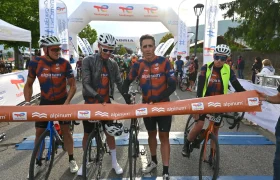 Cuatro ciclistas con camisetas Alpinum TotalEnergies de color naranja y azul a juego se encuentran con sus bicicletas en la línea de partida, preparándose para una carrera.