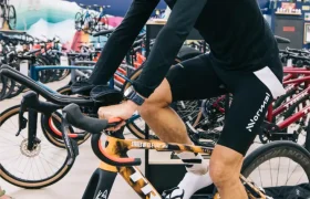 Un hombre vestido con ropa deportiva prueba una bicicleta estática dentro de una tienda de bicicletas, rodeado de equipos de ciclismo, canalizando el espíritu de States of Elevation - Kilian Jornet mientras explora nuevas alturas en el entrenamiento en interiores.