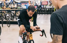 Un hombre monta en una bicicleta estática en una tienda de bicicletas mientras otra persona observa, con varias bicicletas expuestas al fondo, capturando el espíritu de States of Elevation - Kilian Jornet.
