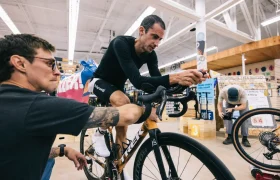 Un hombre con ropa de ciclista se coloca en una bicicleta estática en una tienda, mientras otro ajusta el manillar, captando una concentración y determinación silenciosas que recuerdan a Kilian Jornet en sus estados de mayor elevación.