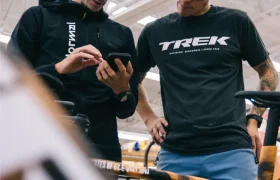 Dos hombres de pie junto a una bicicleta, mirando un smartphone; uno lleva una camiseta "Trek" y el otro una sudadera negra con capucha y gráficos de States of Elevation - Kilian Jornet.