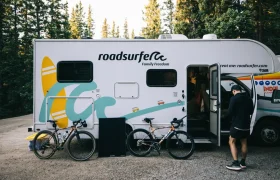 Un hombre junto a dos bicicletas aparcadas delante de una furgoneta camper "roadsurfer" en una zona boscosa, evocando el espíritu de aventura que se encuentra en States of Elevation - Kilian Jornet.