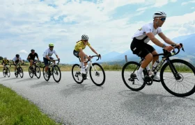 Cinco ciclistas circulan en fila por una carretera rural asfaltada, evocando el espíritu del Tour de Francia, con montañas y un cielo nublado de fondo.