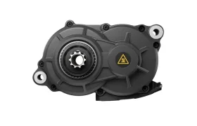 Motor central negro Avinox M2S para bicicletas eléctricas con un símbolo de precaución en el lado derecho, visto desde arriba sobre un fondo blanco.