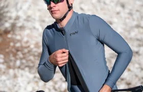 Una persona que lleva gafas de sol y casco se abrocha la cremallera de una chaqueta de ciclista azul con las letras «NW», de pie al aire libre.