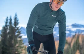 Un ciclista que lleva gafas de sol, guantes y una chaqueta de color verde azulado va en bicicleta al aire libre, con montañas y árboles al fondo.