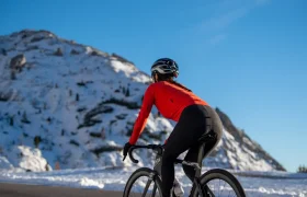 Un ciclista, con una chaqueta roja y casco, circula en bicicleta de carretera por una carretera asfaltada, con una montaña cubierta de nieve al fondo.