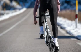 Un ciclista, que lleva mallas negras y zapatillas blancas, va en bicicleta de carretera por una carretera asfaltada con nieve a los lados.
