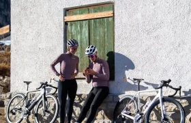 Dos ciclistas, vestidos con el mismo equipo, están junto a una pared con sus bicicletas; uno de ellos mira el móvil y ambos sonríen bajo los cascos y las gafas de sol.