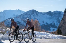 Dos ciclistas pedalean por una carretera asfaltada a través de un paisaje montañoso nevado, con un cielo despejado y picos lejanos al fondo.