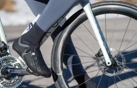 Primer plano de una persona montando en bicicleta, en el que se ve la zapatilla negra de ciclismo del ciclista enganchada al pedal y parte de la rueda delantera y la horquilla de la bicicleta.