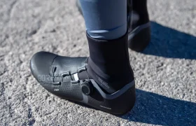 Una persona que lleva unas zapatillas de ciclismo negras con cierre de dial y unos leggings de color gris azulado está de pie sobre una superficie de asfalto irregular.