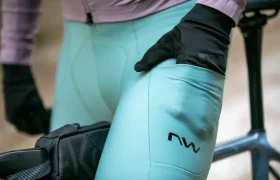 Una persona que lleva unos culotes de ciclismo de color verde azulado y guantes negros está de pie junto a una bicicleta, con una mano apoyada en el muslo. Los culotes tienen un logotipo que dice «nw».