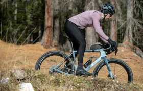 Una persona que lleva casco, guantes y ropa de ciclismo pedalea con una bicicleta de gravel cuesta arriba por un sendero cubierto de hierba, con árboles al fondo.