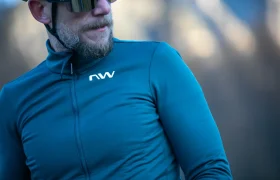 Un hombre que lleva gafas de sol y un maillot de ciclismo azul de manga larga con el logotipo «nw» en blanco está de pie al aire libre.