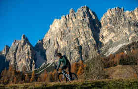 Una persona va en bicicleta por un camino cubierto de hierba, con altas montañas rocosas y un cielo azul y despejado al fondo.