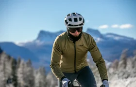 Un ciclista, que lleva casco, gafas de sol y chaqueta, pedalea al aire libre con montañas y árboles al fondo.