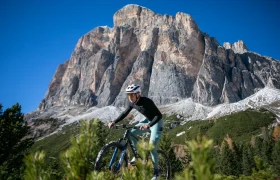 Un ciclista, con casco y gafas de sol, va en bicicleta de montaña por un sendero, con una gran montaña rocosa y un cielo azul y despejado al fondo.