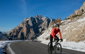 Un ciclista, con casco y chaqueta roja, sube en bicicleta por una carretera de montaña asfaltada, rodeado de nieve y picos rocosos, bajo un cielo azul y despejado.