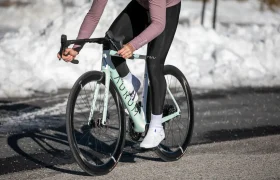Un ciclista, vestido con manga larga y mallas, pedalea en una bicicleta de carretera de color turquesa por una carretera asfaltada con nieve a los lados.