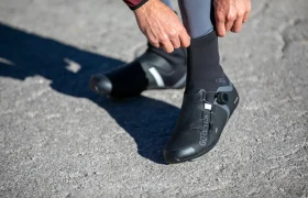 Una persona se coloca unos cubrezapatillas negros de ciclismo sobre las zapatillas mientras está de pie sobre una superficie pavimentada.