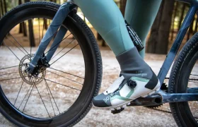 Primer plano de una persona que lleva mallas verdes y zapatillas de ciclismo, pedaleando en una bicicleta por un camino de tierra que atraviesa una zona boscosa.
