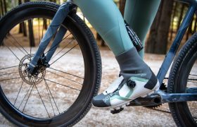 Primer plano del pie de un ciclista con unas zapatillas de ciclismo blancas y verdes enganchadas al pedal de una bicicleta, con un camino de grava y árboles al fondo.