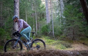 Una persona que lleva casco y gafas de sol va en bicicleta por un sendero de tierra que atraviesa un bosque con árboles altos y frondosa vegetación.