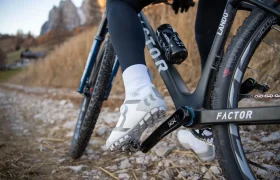Primer plano del pie de un ciclista, calzado con una zapatilla blanca de ciclismo, sobre el pedal de una bicicleta Factor negra, mientras circula por un sendero rocoso.