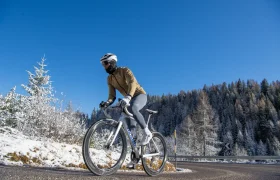 Una persona que lleva casco y ropa de ciclismo de invierno va en bicicleta de carretera por una carretera asfaltada, con árboles cubiertos de nieve y un cielo azul despejado al fondo.