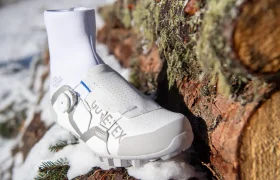 Una zapatilla de ciclismo blanca, con un calcetín alto hasta el tobillo, descansa sobre una pila de troncos cubiertos de una ligera capa de nieve; presenta la palabra «VULCLEX» en el lateral y un sistema de cierre con rueda giratoria.
