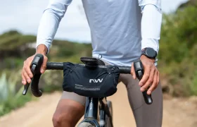 Persona vestida con una camiseta blanca de manga larga y que lleva un reloj, montada en bicicleta por un camino de tierra, con las manos agarradas al manillar, en el que hay una pequeña bolsa negra.