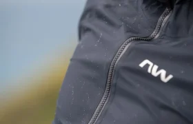 Primer plano de una chaqueta impermeable oscura con gotas de agua en la superficie y un logotipo blanco con las letras «NW» visible cerca de la cremallera.