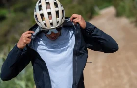 Una persona que lleva casco y gafas de sol se abrocha la cremallera de una chaqueta oscura sobre una camisa blanca mientras está de pie en un camino de tierra al aire libre.
