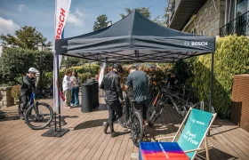 La gente se reúne bajo una carpa con la marca Bosch en la que se exponen bicicletas eléctricas sobre una cubierta de madera, explorando la experiencia sobre la bici con el Bosch Performance Upgrade para más potencia. Cerca hay una tumbona y material promocional.