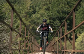 Una persona con casco y gafas de sol conduce una bicicleta de montaña con más potencia, cruzando un estrecho puente metálico rodeado de densos árboles verdes.