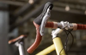 Primer plano de la cinta de manillar marrón de una bicicleta de carretera, la palanca de freno plateada y parte de un cuadro amarillo con grupos de carretera Shimano CUES, sobre un fondo borroso de interior.