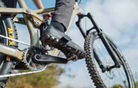 Primer plano del pie de una persona sobre el pedal de una bicicleta de montaña, en el que se aprecian los pedales Shimano DEORE XT SPD XC Gravel Trail, con la cadena, el cambio y la rueda a la vista, al aire libre bajo un cielo parcialmente nublado.